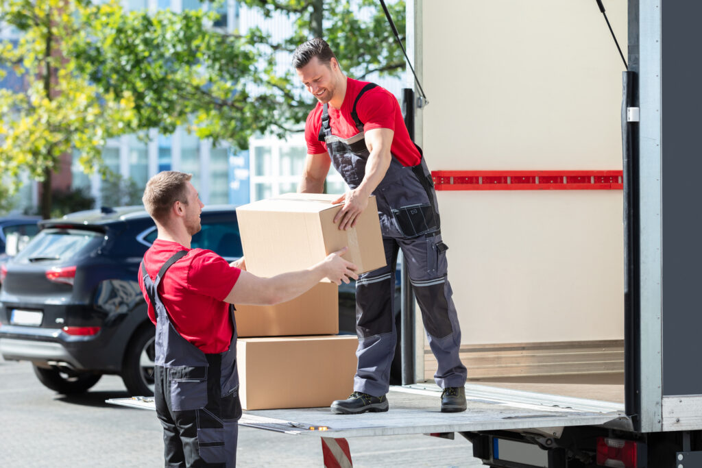 Two Young Male Movers In Uniform Carrying Cardboard Boxes From Truck